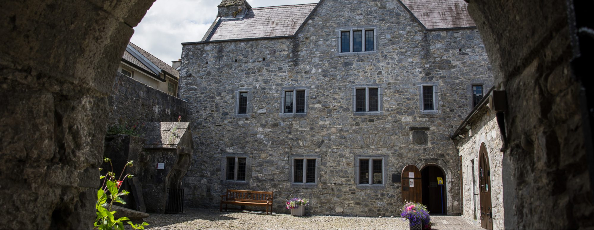 Stone courtyard with a large old building, bench and flower pots, seen through a rocky archway